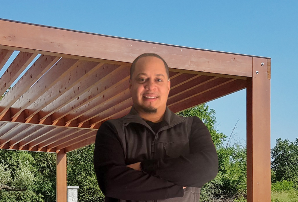 Smiling man with a short beard, wearing a black vest, standing with arms crossed in front of a wooden pergola and lush greenery under a clear blue sky