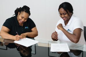 Two women celebrating a successful real estate closing, one signing documents and the other smiling, representing the stress-free home-selling process with We Buy Any House As Is.